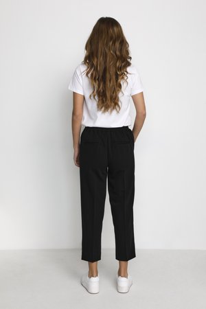 Donna con capelli lunghi e mossi, che indossa una maglietta bianca, pantaloni neri a culotte e sneakers bianche, in piedi di fronte a un muro bianco semplice.