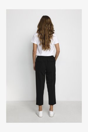 Woman with long wavy hair wearing a white t-shirt, black cropped pants, and white sneakers, standing facing a plain white wall.