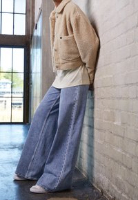 Personne appuyée contre un mur en briques blanches, portant une veste sherpa beige, un jean bleu clair à jambes larges et des baskets blanches à l'intérieur.