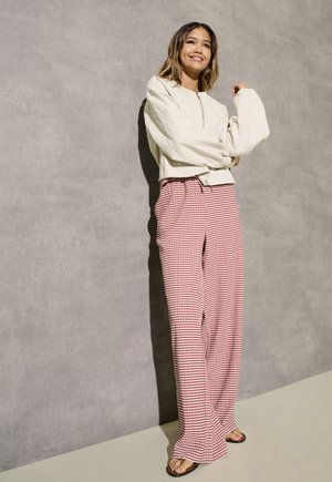 Woman standing against gray wall wearing white textured long-sleeve top, red and white striped wide-leg pants, and black sandals.