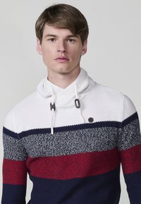 Joven con el cabello corto que lleva un suéter de punto a rayas elegante en blanco, azul marino y rojo, de pie contra un fondo sencillo.