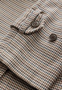 Gros plan sur la manche d'une veste pied-de-poule marron et beige avec deux boutons marron et un détail de lanière en tissu.
