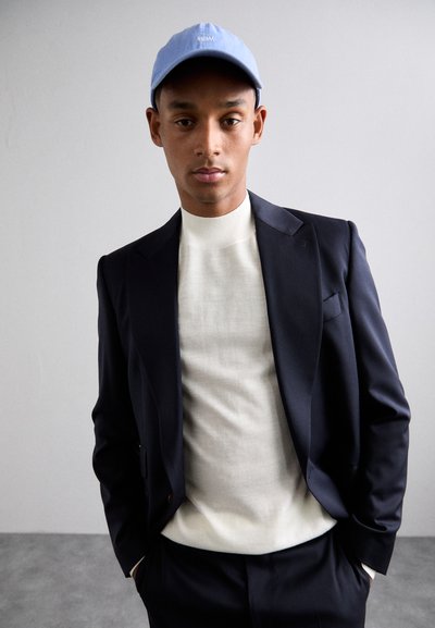 Uomo che indossa un berretto azzurro chiaro, un maglione a collo alto bianco e un giubbotto e pantaloni blu navy scuro, in piedi di fronte a uno sfondo grigio uniforme.