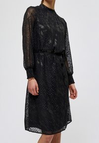 Černé šaty s průhlednými dlouhými rukávy, texturovaným vzorem, nařaseným pasem a vysokým límcem. Doplňky zahrnují nařasené manžety a splývavý střih.