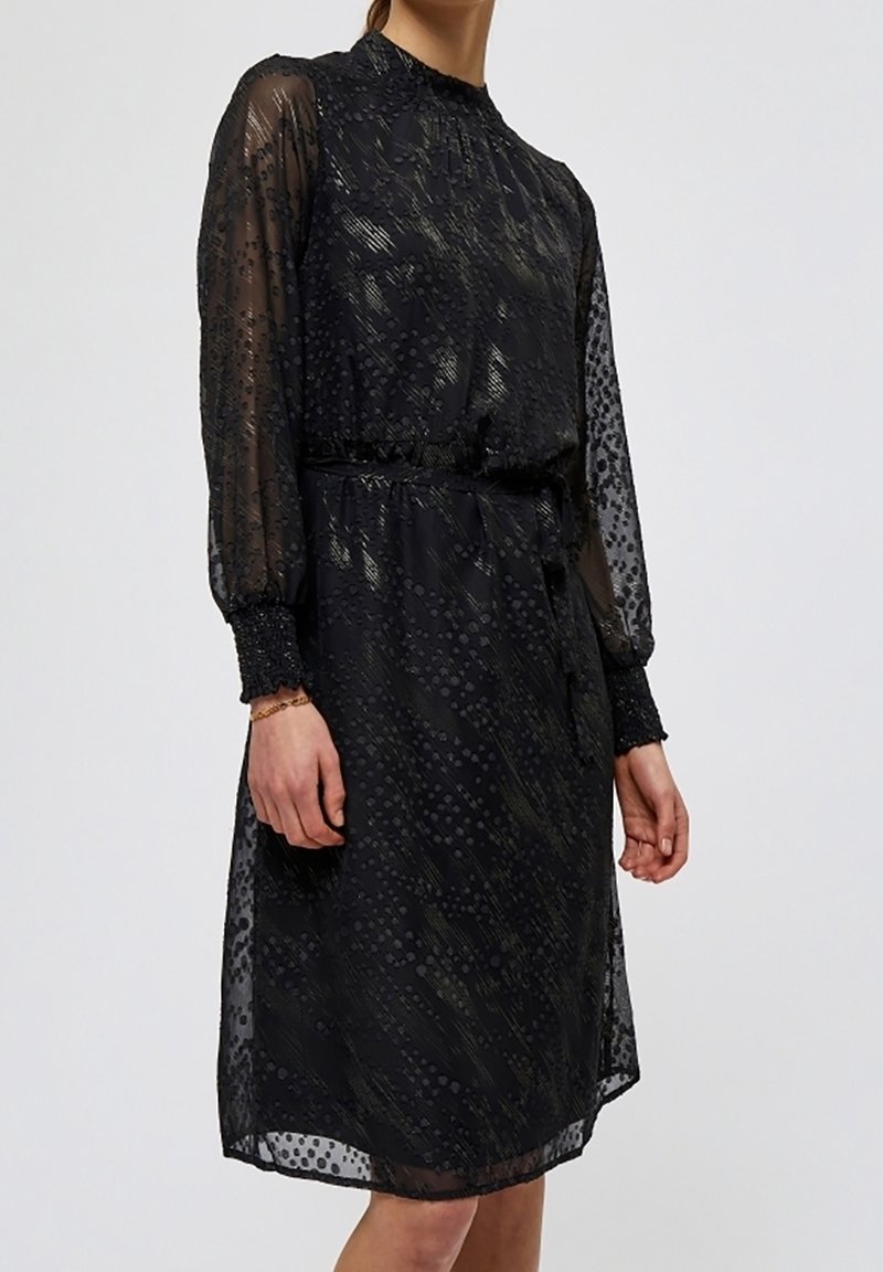 Černé šaty s průhlednými dlouhými rukávy, texturovaným vzorem, nařaseným pasem a vysokým límcem. Doplňky zahrnují nařasené manžety a splývavý střih.