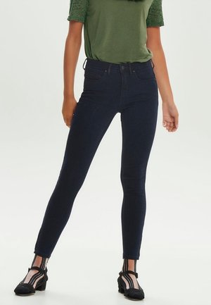 Femme portant un jean skinny bleu foncé, un haut vert à manches courtes avec des manches en dentelle, et des sandales à talons noires avec des chaussettes transparentes rayées.