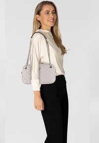 Lichtgrijze, gewatteerde handtas met een rechthoekige vorm, zilveren kettingriem en subtiele hardwareaccenten. Gestyled met een crème blouse en zwarte broek.