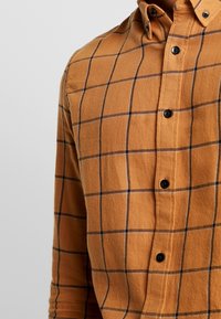 Camicia con bottoni di colore arancione ruggine, realizzata in tessuto testurizzato. Presenta un motivo a quadri neri, maniche lunghe e colletto arrotondato con bottoni neri.
