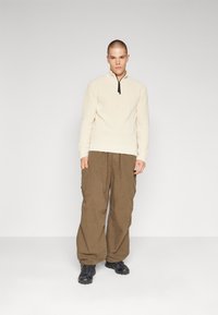 Suéter de punto beige con cuello alto y cremallera negra, combinado con pantalones cargo sueltos de color marrón oscuro y zapatillas negras.
