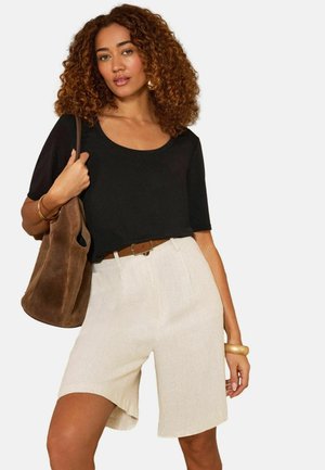 Kurdade juustega naine, kes kannab musta sügava kaelusega t-särki, beežika värvi kõrge vöökohaga lühikesi pükse, pruuni vööd, pruuni õlakotti ja kuldseid ehteid.