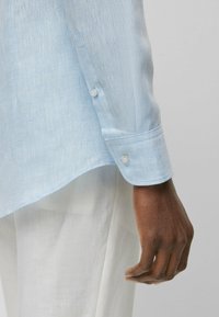 Camicia di lino azzurro chiaro con maniche lunghe, dotata di un bottone al polsino e di un orlo curvo, esposta accanto a un tessuto bianco.