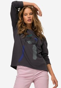 Sudadera gris oscuro de manga larga, de corte holgado, con diseños gráficos abstractos en azul y verde. Combinada con pantalones rosas claros.