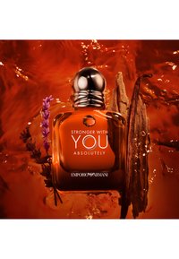 Botella de perfume Emporio Armani etiquetada "Stronger With You Absolutely" rodeada de vainas de vainilla y lavanda con un fondo de líquido ámbar.