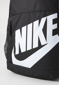Juodas kuprinės modelis su dideliu baltu Nike logotipu, pagamintas iš patvarios medžiagos, turi šoninį tinklinį kišenę ir užtrauktuko skyrių.