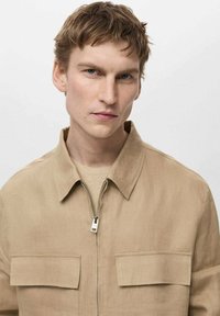 Beige linned zip-up skjorte med krave, to forlommer og et rent, minimalistisk design. Har en glat tekstur og enkelt hardware.