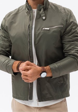 Uomo che indossa una giacca verde oliva con zip sopra una camicia bianca e pantaloni beige, un orologio da polso e braccialetti con perline di legno, mani giunte davanti.