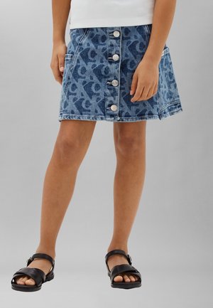 Copil purtând o fustă din denim albastru cu model și nasturi în față, un top alb și sandale negre cu bretele, stând pe fundal gri.