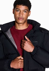 Schwarz gepolsterte Jacke mit hohem Kragen, ausgestattet mit einem grauen Futter und einer kleinen schwarzen Akzenttasche, getragen über einem burgunderrot strukturierten Hemd.