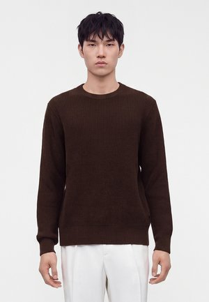 Jeune homme aux cheveux courts et foncés portant un pull côtelé marron foncé et un pantalon blanc, debout devant un fond clair uni.