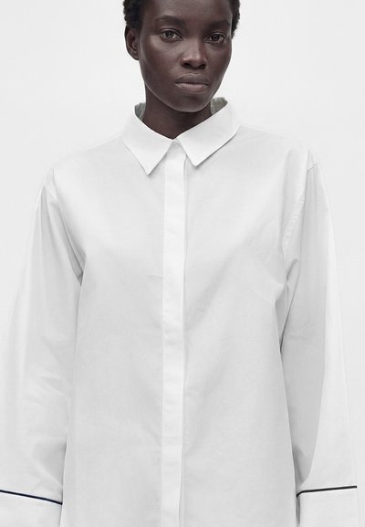 Personne à la peau foncée portant une chemise blanche oversize boutonnée avec un col pointu et un liseré noir aux poignets, fond uni.