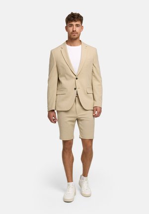 Mand stående iført beige blazer og matchende shorts med hvid t-shirt og hvide sneakers mod ensfarvet baggrund.