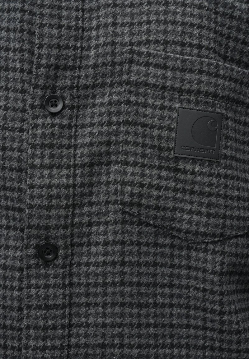 Graues Flanellhemd mit einem Hahnent pattern, ausgestattet mit schwarzen Knöpfen und einem quadratischen schwarzen Logo-Patch auf der Brusttasche.