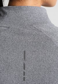 Graues Sportshirt mit hohem Kragen, aus strukturiertem Stoff und mit vertikalem Logodetail auf dem Rücken. Sichtbare, glatte Nähte.