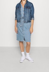 Farkkutakki kevyen sinisen farkkuompeluksen päällä, jossa on narulla säädettävä vyötärö, napitettava etuosa ja sivuhalkiot, yhdistettynä valkoisiin tennareihin.