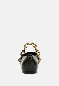 Zapato plano de cuero negro con punta redondeada y una correa de cadena dorada. Presenta un pequeño logo en la parte trasera. Textura suave, diseño minimalista.