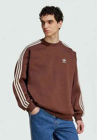 Joven con un sudadera marrón de Adidas con rayas blancas en las mangas y jeans azules, mirando hacia un lado con una mano en el bolsillo.