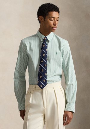 Άνδρας που φοράει πράσινο ριγωτό πουκάμισο με κουμπιά, σκούρο μπλε γραβάτα με σχέδια ασπίδας και κρεμ πλεκτό παντελόνι με πιέτες ψηλόμεσο, μπροστά σε ουδέτερο φόντο.