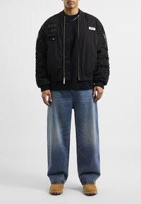 Giacca bomber nera con texture trapuntata e accenti del logo, abbinata a jeans a gamba larga blu chiaro e stivaletti beige.