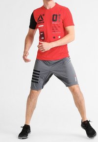 Camiseta atlética de manga corta en rojo y negro con gráficos, combinada con pantalones cortos grises con rayas negras. Unos tenis completan el atuendo.