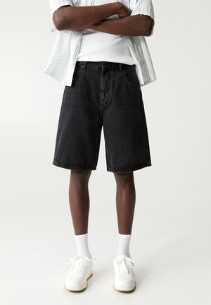 Personne portant un short en jean noir jusqu'aux genoux, des chaussettes blanches, des baskets blanches, un débardeur blanc et une chemise à manches courtes rayée déboutonnée.
