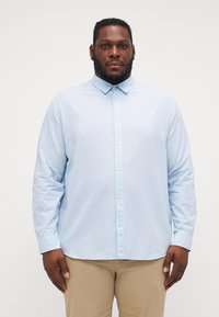 Chemise longue à manches longues bleu clair avec un col boutonné et des boutons à l'avant, fabriquée en tissu texturé. Présente une coupe décontractée et des manches à poignets.