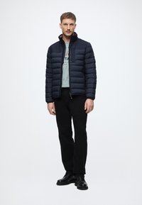 Tengerész kék puffertakó, párnázott kialakítással, magas gallérral és oldalsó zsebekkel; világosszürke ing felett viselve, fekete nadrággal és csizmával párosítva.