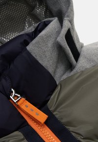Gewatteerde jas met grijs fleece, marineblauwe nylon en olijfgroene stof. Inclusief een oranje ritssluiting en zwarte accentstrip. Gestructureerde binnenkant.