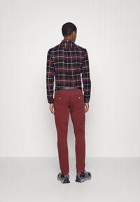 En man står med ryggen mot kameran, iklädd en röd och svart rutiga skjorta och vinröda byxor, tillsammans med grå sneakers.