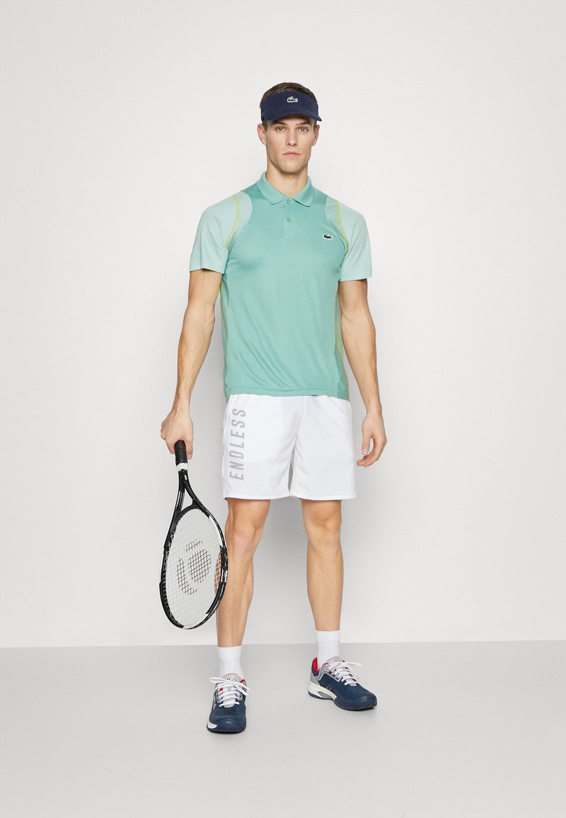 Homme portant un polo vert menthe, un short blanc avec le texte "ENDLESS", une visière marine et des baskets, tenant une raquette de tennis et adossé à un fond uni.