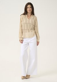 Beige lace blouse with a V-neck, long sleeves, and intricate patterns; paired with wide white trousers and beige heeled shoes.