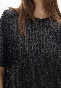 Svart paljett-topp med en löst sittande passform, korta ärmar och rund halsringning. Texturen är slät med ett skimrande mönster över hela.