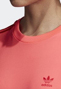 Kortärmad t-shirt med rund halsringning i ljus korallfärgad tyg. Har en ribbad halsringning och en liten röd Adidas-logotyp på bröstet. Slät textur.