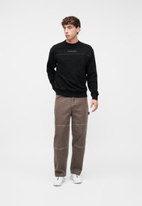 Νεαρός άνδρας όρθιος, φοράει μαύρο φούτερ με το λογότυπο "NAPAPIJRI", καφέ παντελόνι με ραφές και λευκά αθλητικά παπούτσια, κοιτώντας προς τα αριστερά του.