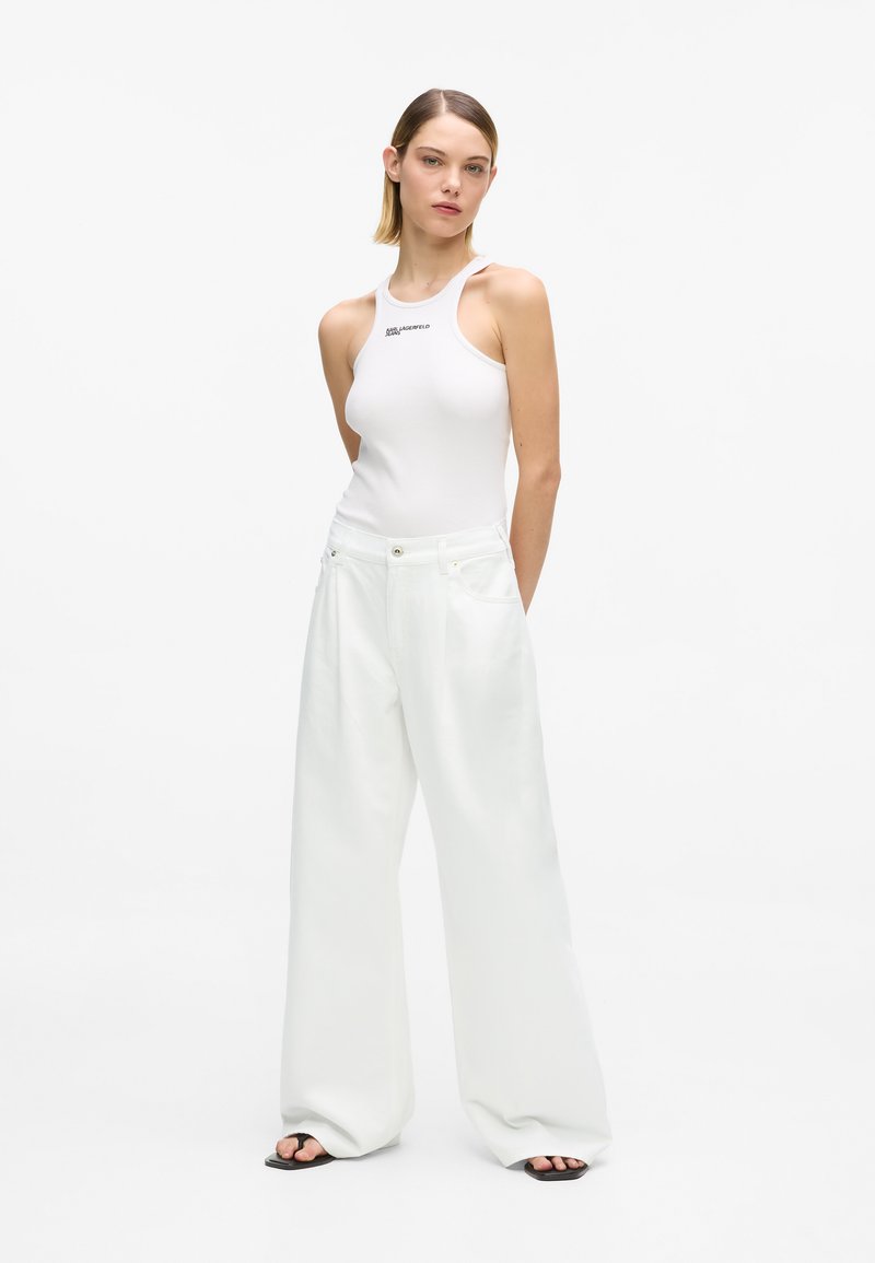 Γυναίκα με λευκό αμάνικο τοπ και λευκό παντελόνι με φαρδιά πόδια, στέκεται με τα χέρια πίσω από την πλάτη σε απλό λευκό φόντο.