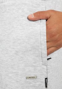 Gray sweatshirt fabric with a textured finish. Features a side pocket and branded tag. Hand resting on the pocket.