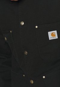 Sort arbejdsjakke lavet af robust stof, med guldtonede trykknapper og to frontlommer med et hvidt og orange logo-patch.