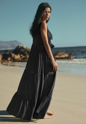 Femeie cu părul lung și închis la culoare, purtând o rochie neagră vaporoasă, stând desculță pe o plajă cu nisip, cu stânci și mare în fundal.