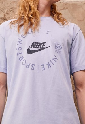 Jasnofioletowa bawełniana koszulka z czarnym logo Nike oraz nadrukowanymi grafikami, z okrągłym dekoltem, krótkimi rękawami i luźnym krojem.