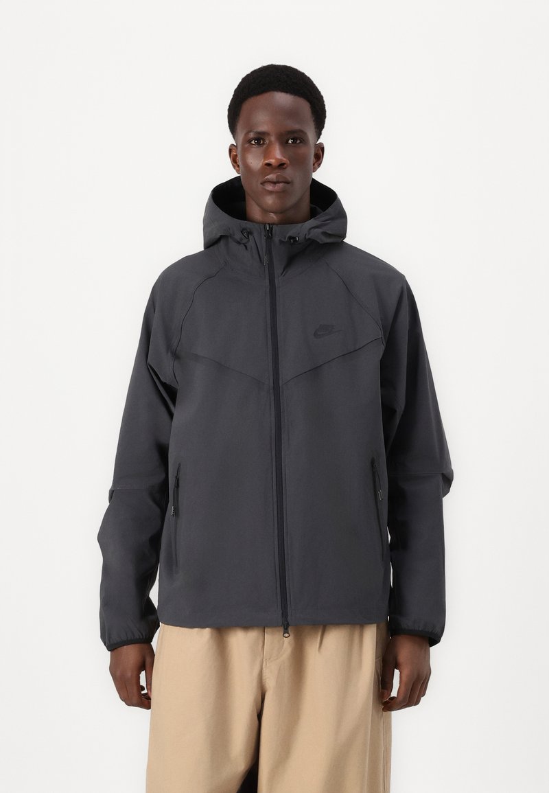 Dunne, donkergrijze hooded jas met een ritssluiting, verstelbare capuchon en twee zijzakken; gladde textuur met een modern ontwerp.