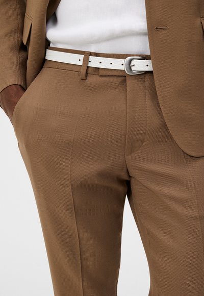 Braune strukturierte Anzughose mit einem weißen Gürtel und einer silbernen Schnalle. Sichtbare Taschen, kombiniert mit einem weißen Unterhemd.